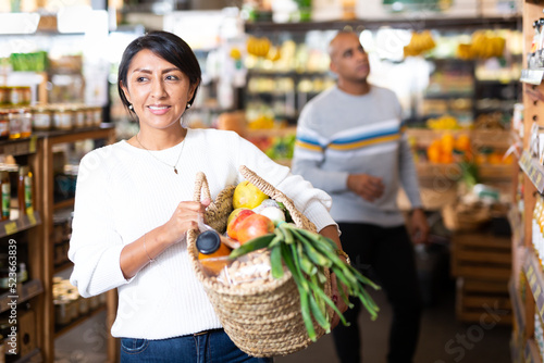 Portrait of smiling latin american woman with wicker basket with fresh grocery, enjoying shopping in store