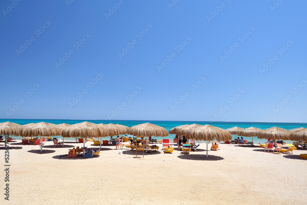 Straw shade umbrellas on sea tropical beach with resting sunbeds against blue vibrant sky in summer
