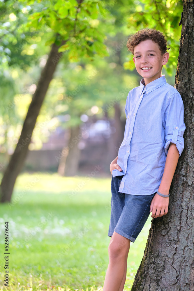 Young happy child boy relaxing in summer park leaning on tree trunk. Positive youngster enjoying summertime outdoors. Child wellbeing concept