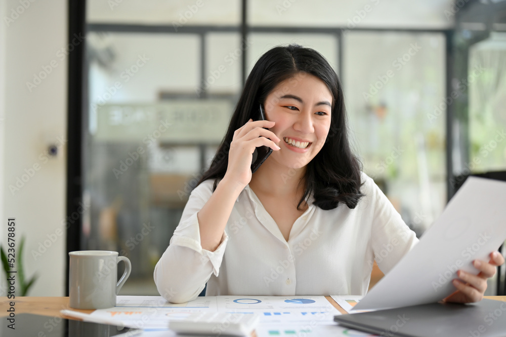 Asian young businesswoman talking on the phone while looking the details on her report. Asian young businesswoman talking on the phone while looking the details on her report.