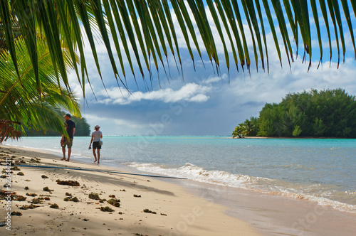 A couple walks barefoot along a tropical beach. viewed from under coconut palm leaf. Rarotonga, Cook Islands