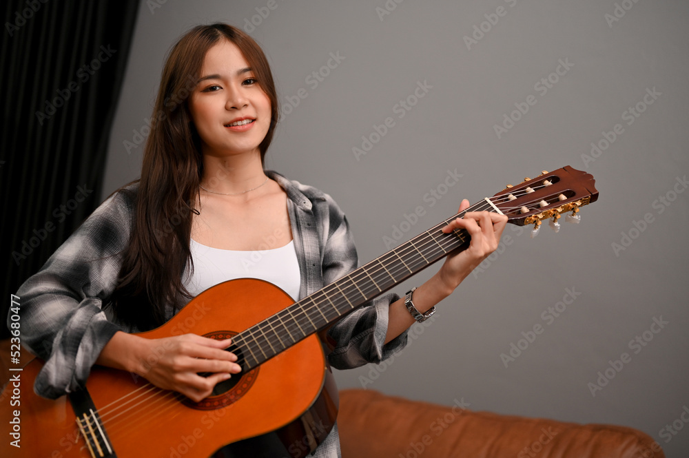 Beautiful young Asian female artist playing an acoustic guitar in her home living room. Beautiful young Asian female artist playing an acoustic guitar in her home living room.