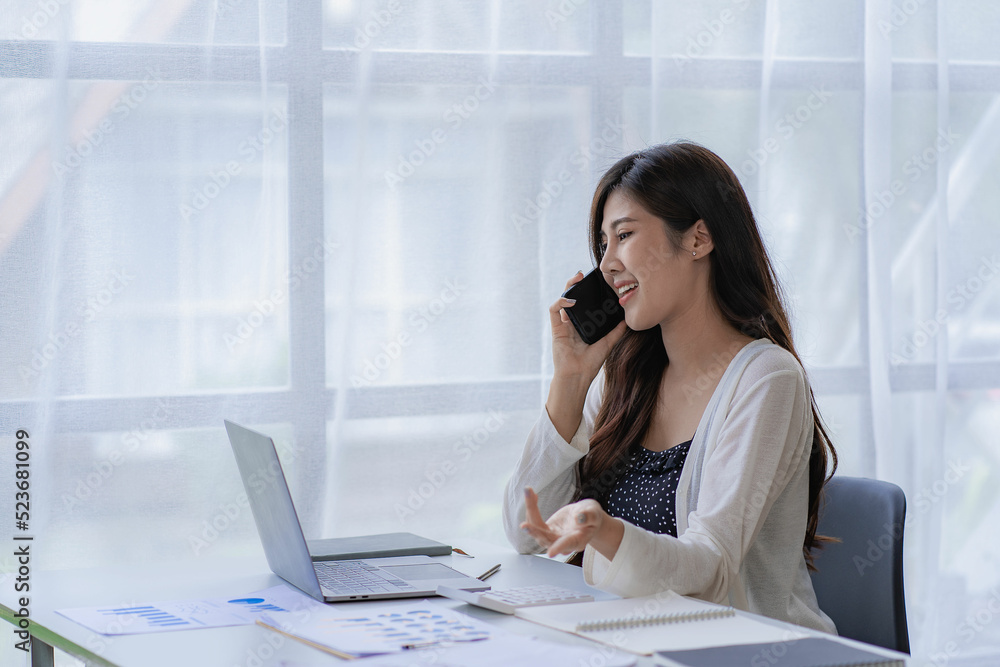 Asian businesswoman using smartphone to do accounting work with laptop and graph of financial reports in office Asian businesswoman using smartphone to do accounting work with laptop and graph of financial reports in office