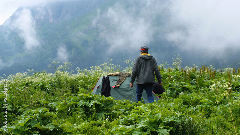 A male hiker walks through tall grass to a tent on which things are drying. In the background of the tent are beautiful mountains obscured by fog.