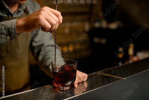 hand of bartender hold long bar spoon and stirring cold drink in glass on bar.
