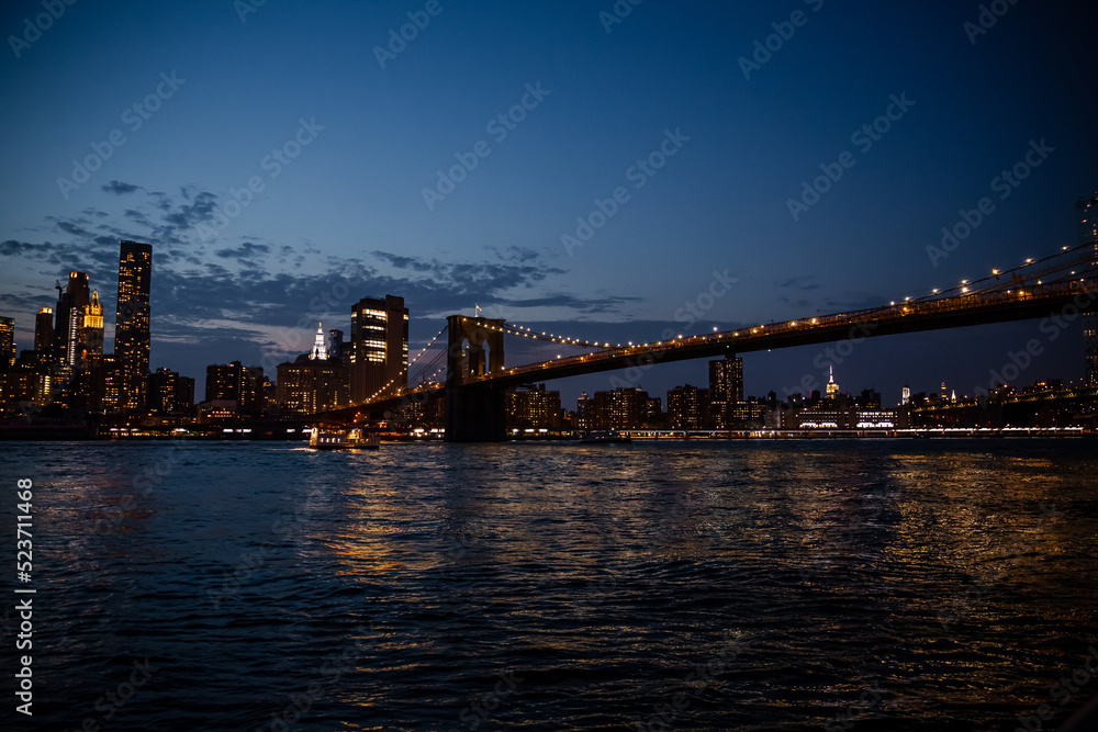Brooklyn bridge with water reflection, New York City Skyline. Beautiful sunset over manhattan with manhattan and brooklyn bridge. Illuminated buildings and brooklyn bridge against sky at night