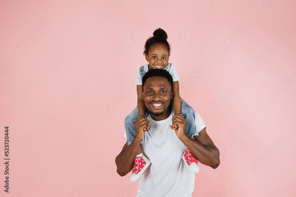 Happy family. Portrait of cheerful african american man riding excited daughter on his shoulders, looking and posing at camera, having fun, holding hands isolated over pink studio background. Happy family. Portrait of cheerful african american man riding excited daughter on his shoulders, looking and posing at camera, having fun, holding hands isolated over pink studio background.
