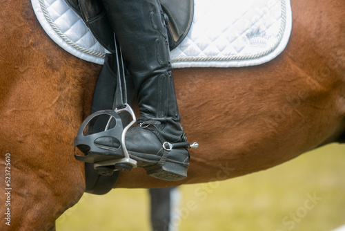 Close up image of jockey riding boot in the stirrup. Rider on bay sport horse.