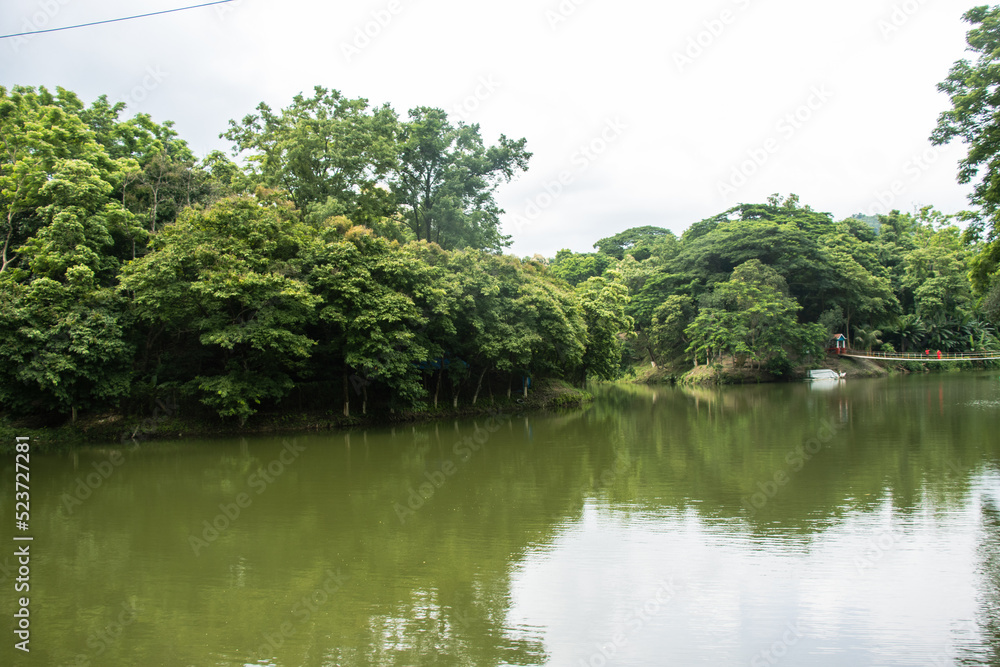 Beautiful rural river flow with tropical forest area. River flow photograph with green trees and cloudy sky. Village river flow view with resort building and bridge. Water Flow photo with hilly forest