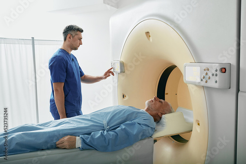 Radiographer operating computed tomography scanner being used to scan lying patient in radiology department of hospital. CT scanner