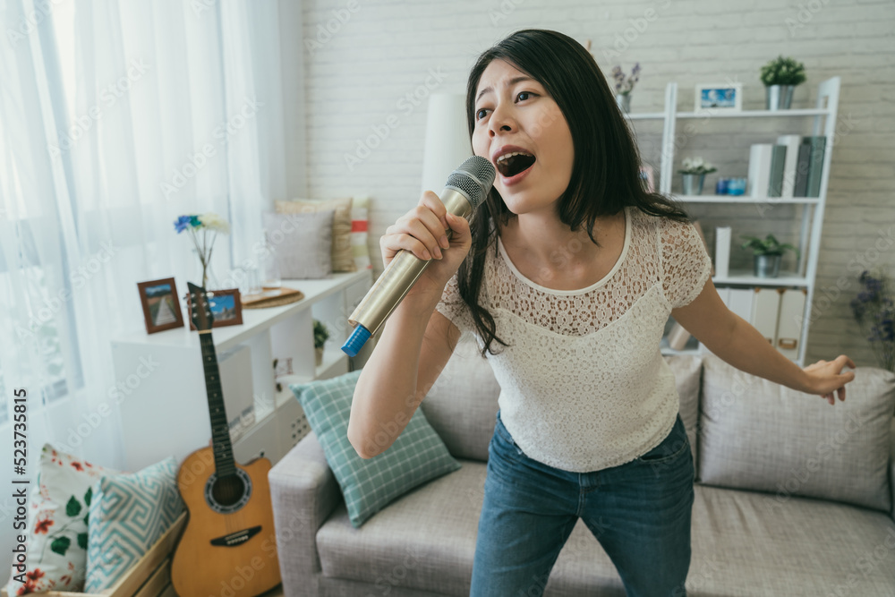 closeup of asian woman holding a microphone and leaning forward while singing karaoke alone loudly full of emotion in the living room at home during daytime. closeup of asian woman holding a microphone and leaning forward while singing karaoke alone loudly full of emotion in the living room at home during daytime.