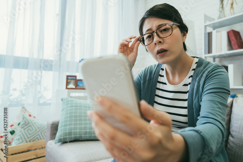 selective focus asian woman having presbyopia is holding her glasses and squinting her eyes while trying to read clearly on the phone at a home living room photo