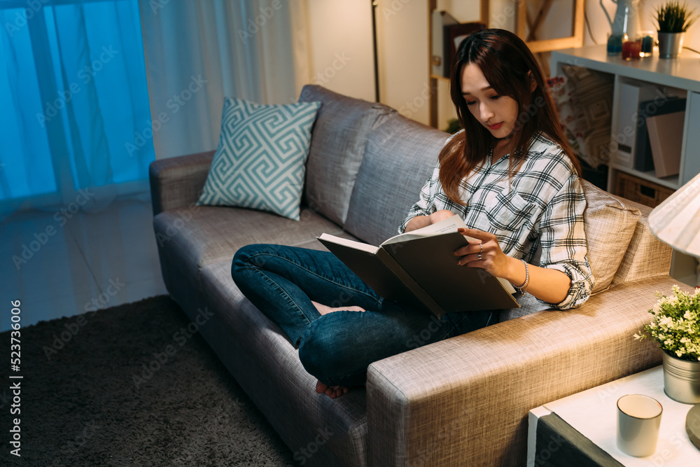 beautiful chinese lady sitting comfortably on the couch is holding and reading a book alone on a peaceful night in a cozy home living room interior. beautiful chinese lady sitting comfortably on the couch is holding and reading a book alone on a peaceful night in a cozy home living room interior.