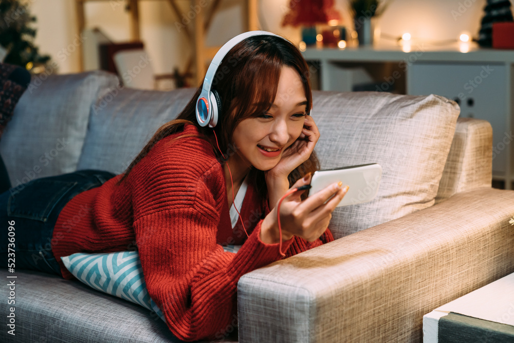 © PR Image Factory - carefree asian girl lying prone on sofa and propping her face is enjoying watching shows through streaming service on her phone in a comfortable home interior.