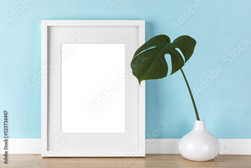Elegant vertical white picture frame with matte mockup, transparent template with monstera leaf in vase in front of pastel blue wall.