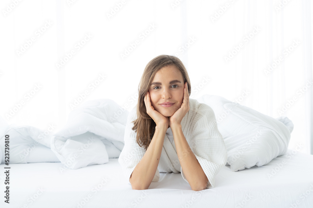 A woman lying at the end of the bed underneath the quilt and smiling, with her head resting upon her hand in the morning A woman lying at the end of the bed underneath the quilt and smiling, with her head resting upon her hand in the morning