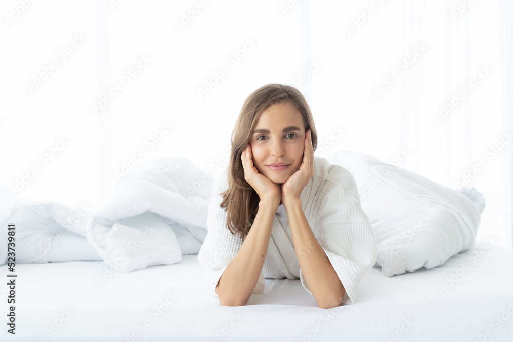 A woman lying at the end of the bed underneath the quilt and smiling, with her head resting upon her hand in the morning A woman lying at the end of the bed underneath the quilt and smiling, with her head resting upon her hand in the morning