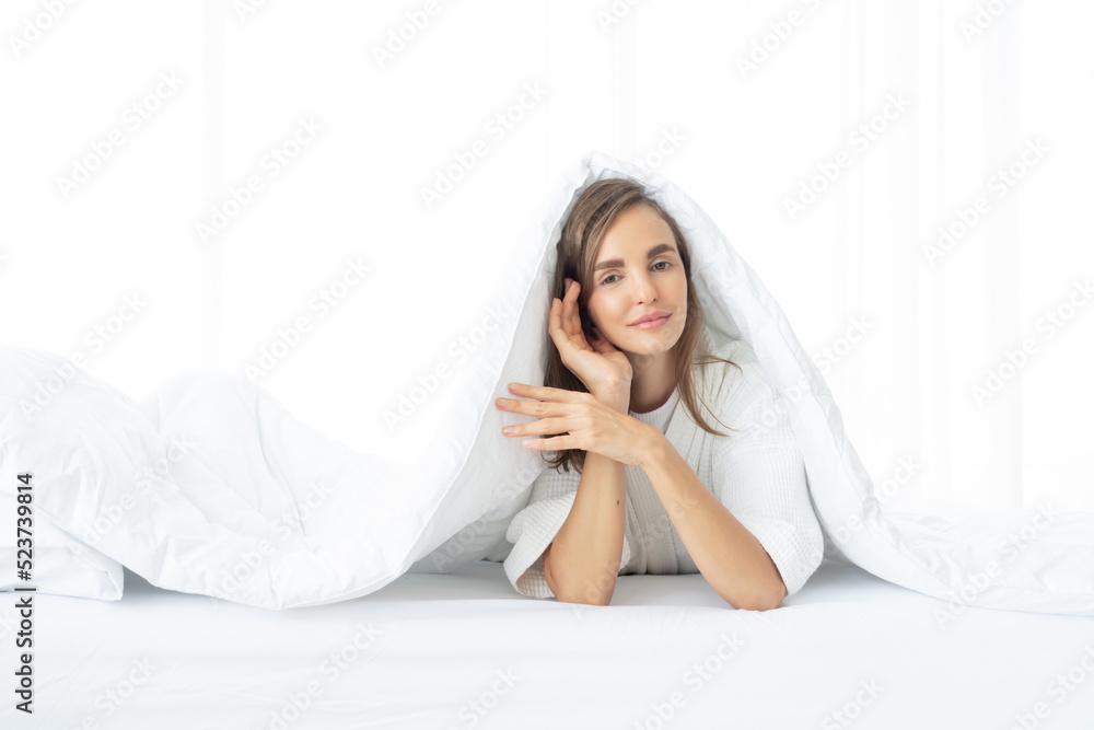 Fototapeta premium A woman lying at the end of the bed underneath the quilt and smiling, with her head resting upon her hand in the morning