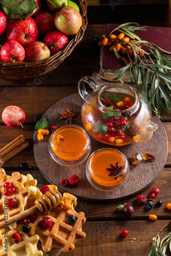 Autumn tea with berries and honey. Sea buckthorn and raspberries in a glass teapot, cups with poured tea. Apples and waffles with honey.