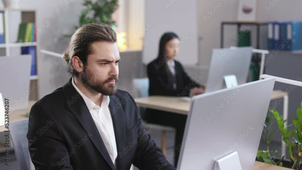 Bearded caucasian man wearing formal suit typing on computer keyboard at modern office with colleague on background. Young male preparing financial diagrams for business presentation. Bearded caucasian man wearing formal suit typing on computer keyboard at modern office with colleague on background. Young male preparing financial diagrams for business presentation.