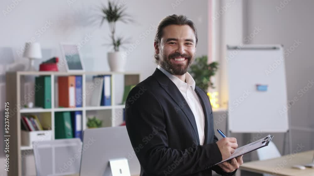 Portrait of bearded man in formal suit holding clipboard and looking at camera with smile. Male worker with long hair making notes before business meeting at office. Portrait of bearded man in formal suit holding clipboard and looking at camera with smile. Male worker with long hair making notes before business meeting at office.