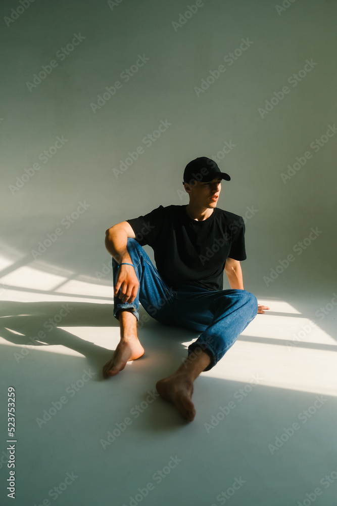 Portrait of stylish handsome caucasian man fashion model in t shirt and cap sitting on a floor, studio shot.