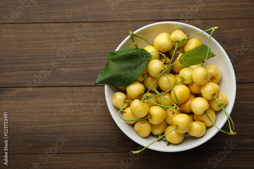 Bowl with ripe yellow cherries and green leaves on wooden table, top view. Space for text