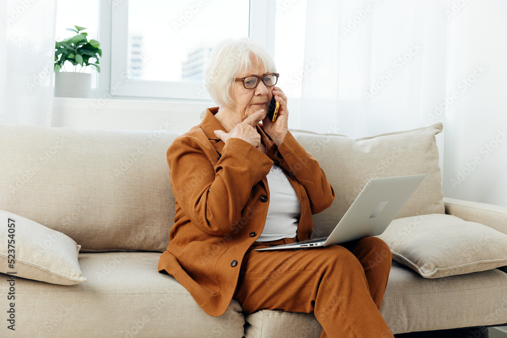 a lovely elderly woman is sitting on a wide beige sofa talking on a smartphone holding a laptop on her lap in a stylish brown suit a lovely elderly woman is sitting on a wide beige sofa talking on a smartphone holding a laptop on her lap in a stylish brown suit