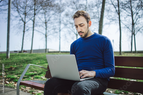 Young bearded man teleworks at his computer in a park sitting on a bench because the weather is nice.