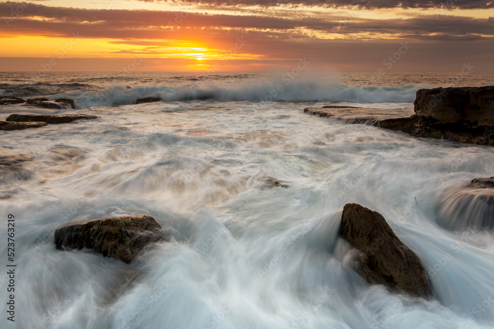 Incoming waves surge over rocks with a golden orange sunrise Incoming waves surge over rocks with a golden orange sunrise