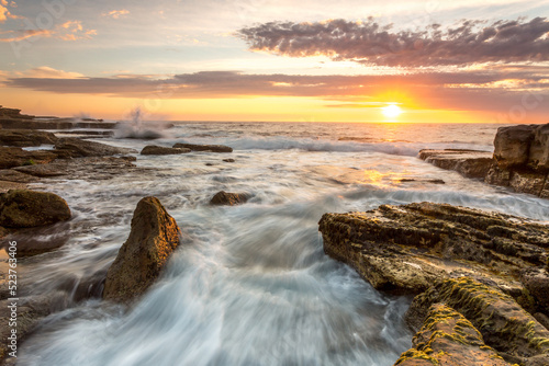 Beautiful Coastal sunrise scene with sun rising over the horizon with foreground waves flowing over rocks