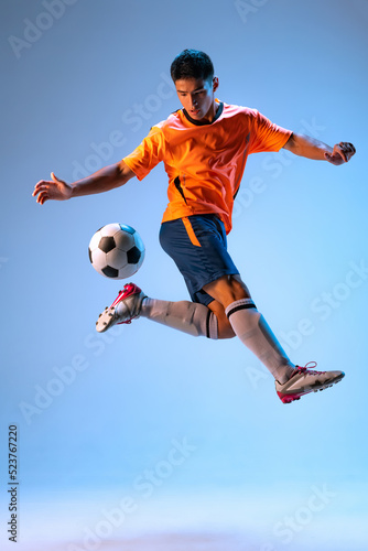 Portrait of young man, football player in motion, training, kicking ball in a jump isolated over blue studio background in neon light © Lustre