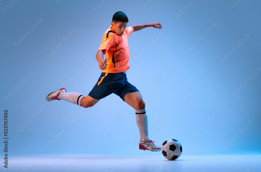 Dynamic portrait of young man, professional football player in motion, training, dribbling ball isolated over blue studio background in neon light