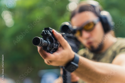 Man wearing camouflage t-shirt safety headphones and goggles aiming machine gun. Outdoor shooting range target practise. Horizontal shot. High quality photo