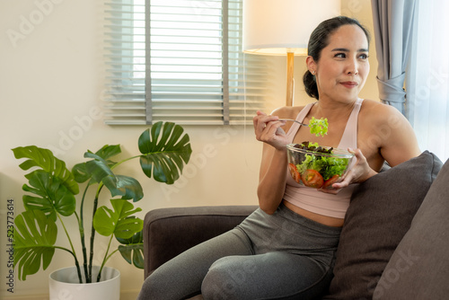 Happy women hold a salad bolw while sitting on a sofa in a living room.