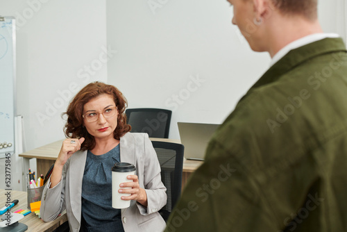Skeptical mature businesswoman drinking coffee and listening to ideas of young employee