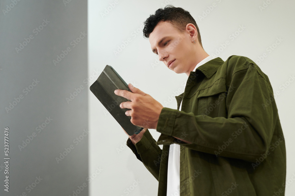 Young man in green denim jacket reading students book on tablet computer Young man in green denim jacket reading students book on tablet computer