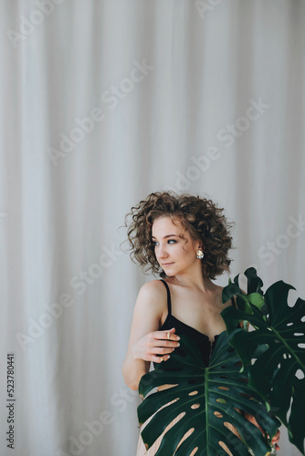 Portrait of a young beautiful woman with blond curly hair holding large green leaves in her hands. Young girl in a black top on a light neutral background
