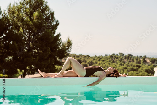 Young beautiful readhead woman laying on the edge of a fantastic swimming pool with a brown swimsuite in the nature