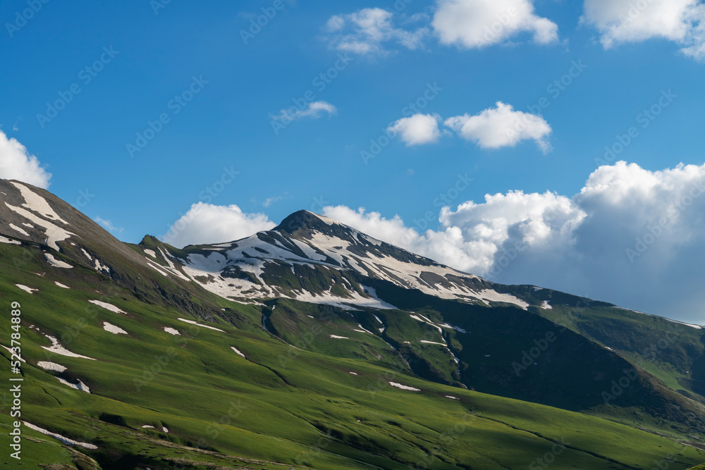 Beautiful snow covered mountains with green meadows under blue sky and clouds. Mountains landscape.