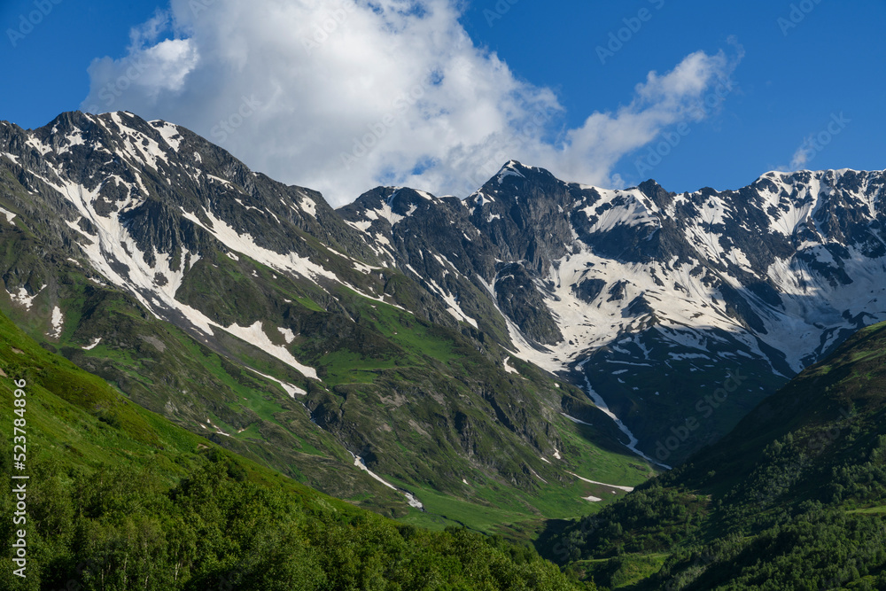 Fototapeta premium Beautiful snow covered mountains with green meadows under blue sky and clouds. Mountains landscape.
