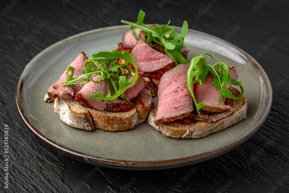 Healthy nutritious breakfast of bruschetta with pastrami beef ham and herbs in a ceramic plate on a dark textured background. Restaurant menu Isolated on black