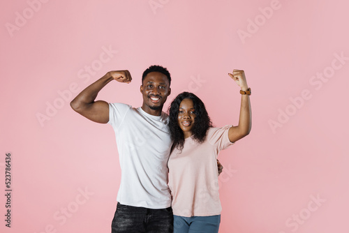 Young cheerful strong sporty fitness african couple two friends man woman in casual t-shirts showing biceps muscles on hand isolated on pastel pink color background studio portrait.