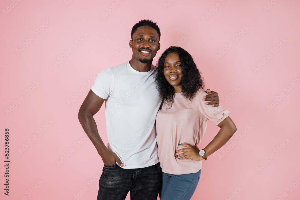 Photo below belt of cheerful people of exotic appearance on isolated nacre pink wall. African female with her arms on waist dressed in pink T-shirt and jeans, while guy in white t-shirt. Photo below belt of cheerful people of exotic appearance on isolated nacre pink wall. African female with her arms on waist dressed in pink T-shirt and jeans, while guy in white t-shirt.