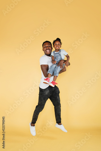 Full length shot of jumping african father holding cute little daughter having fun together. Studio photo of young family on yellow background, copy space.