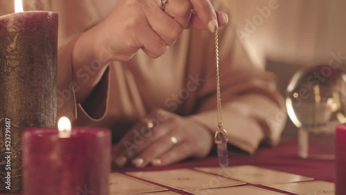 Cropped slowmo closeup of unrecognizable jeweled fortune teller holding pendulum over tarot cards spread on table photo