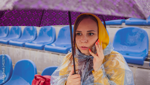 A beautiful woman in a hoodie and a plastic raincoat, talking on a mobile phone, sitting on the podium with an umbrella in her hand during the rain