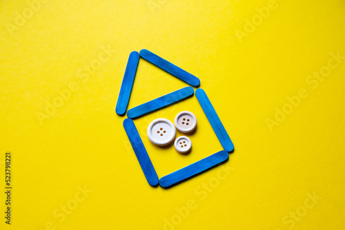 House made of blue sticks on a yellow background. Wooden buttons as a symbol of the family. Housing for Ukrainians. Ukraine flag colors