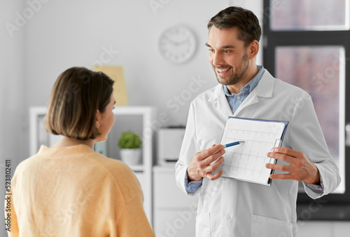 medicine, healthcare and people concept - smiling male doctor or cardiologist with clipboard showing cardiogram to woman patient at hospital