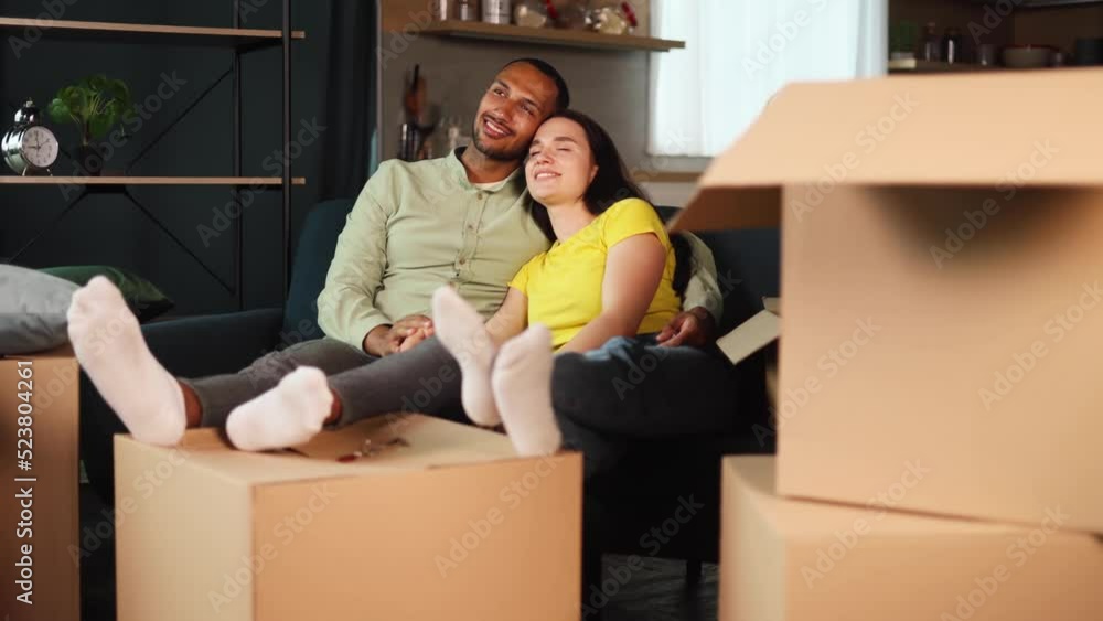 Happy interracial couple sitting on a living room of their new home with a lot of boxes in the background. Married couple plan apartment decoration and imagine their beautiful life. Happy interracial couple sitting on a living room of their new home with a lot of boxes in the background. Married couple plan apartment decoration and imagine their beautiful life.